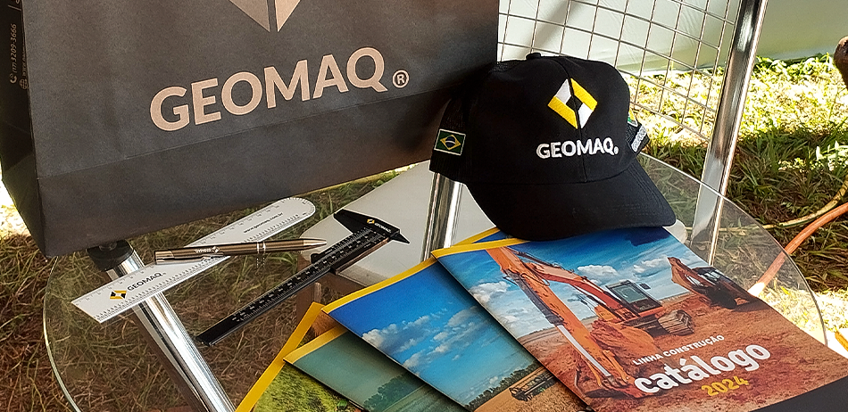 Soluções técnicas e materiais promocionais da Geomaq apresentados na Agrishow 2025, incluindo boné, catálogos e brindes sobre mesa de vidro.